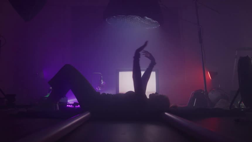 A woman laying on her back gently swayinh her arms in the air in the background is a tube television that has an unstable picture playing on it the room has red and purple mood lighting