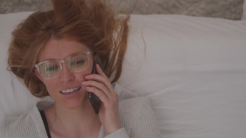 Woman with red hair wearing glasses laying on edge of bed talking on cell phone