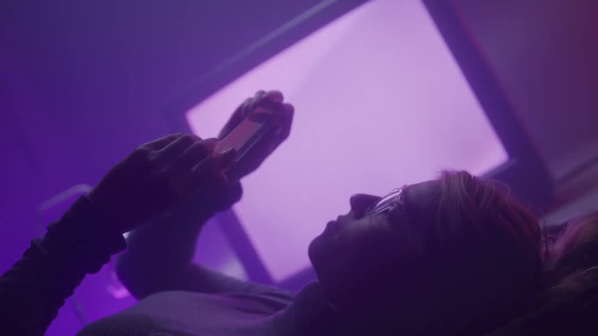 Woman examines a triangular clear crystal while lying down with the television flashing beside her