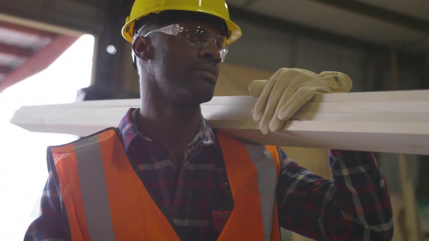 A construction worker wearing a hard hat carries lumber across a workspace