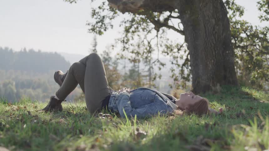 A woman with light brown hair lies on her back in the grass eyes closed wearing a denim jacket and pants with a large tree casting a shadow over her and a scenic landscape in the background
