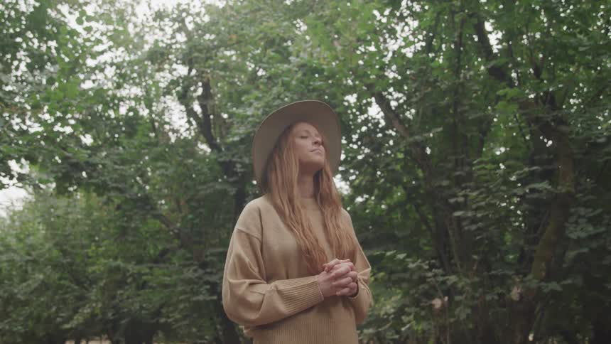 A woman with long light brown hair wearing a hat and a beige sweater stands with her hands clasped eyes closed in a wooded area with tall trees