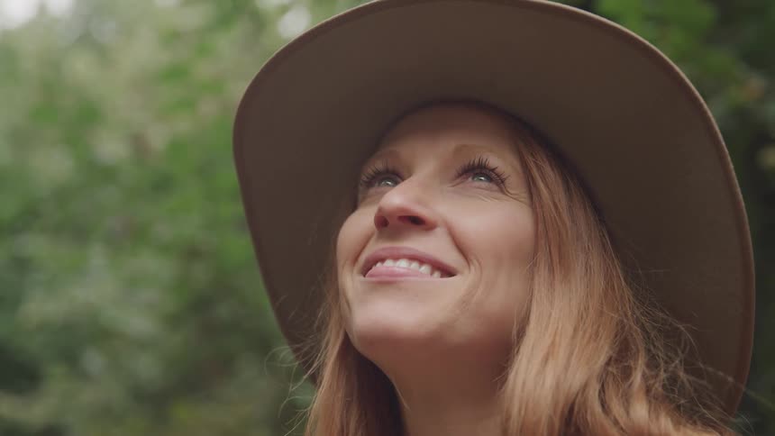 A blonde woman wearing a sun hat is looking up at the tree canopy and smiling