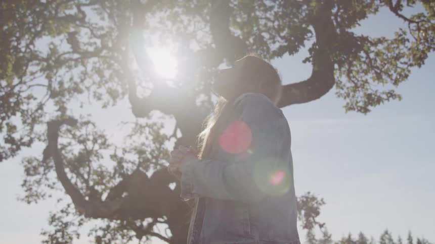 A woman with long light brown hair stands looking up towards the bright sunlight filtering through the branches of a large tree