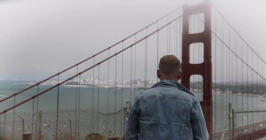 A man approaches and stands before a fence and looks at a view of the golden gate bridge