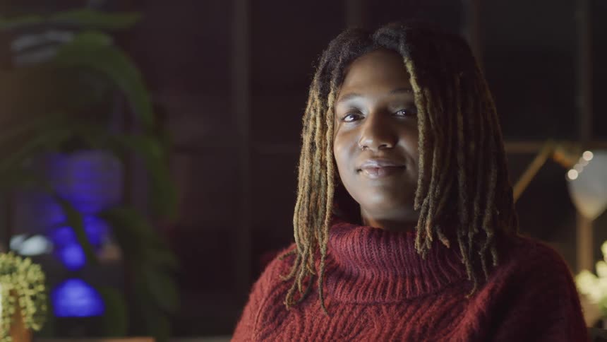 A woman with dreadlocks sits in a study looking at the camera and smiling