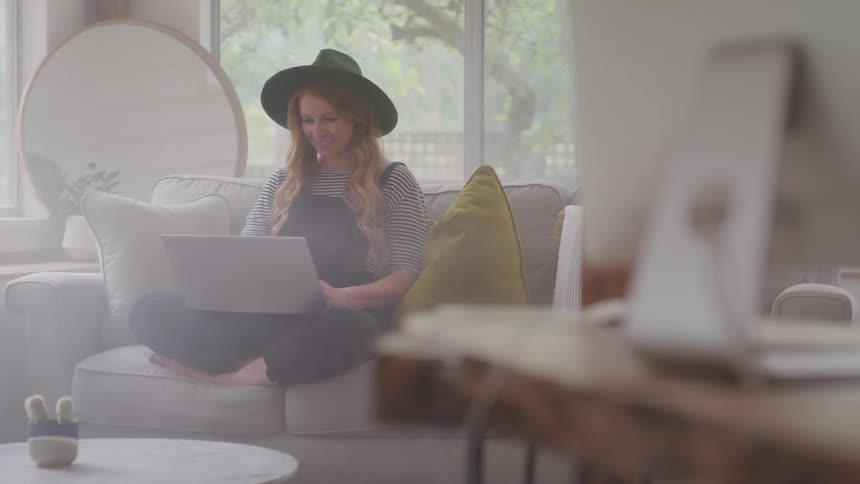 Woman sits on the couch and smiles while typing on a laptop