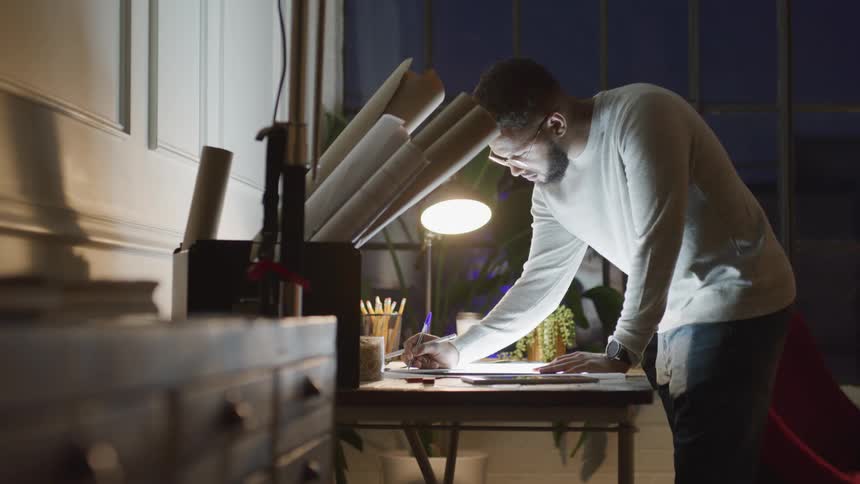 Man standing and drawing at a desk with rolled up plans nearby