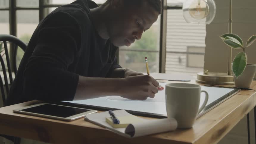 A focused man works on a drawing at a brightly lit table with a pencil and various supplies