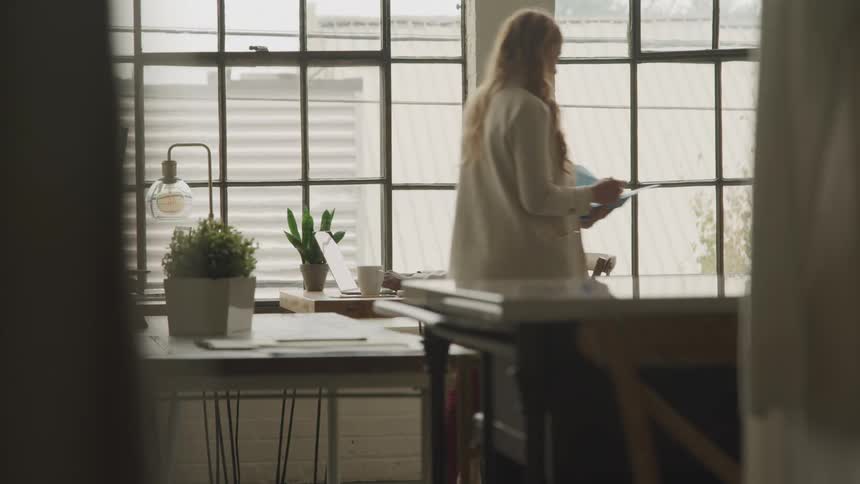 A man works on a laptop near large windows in a bright office as a woman walks past