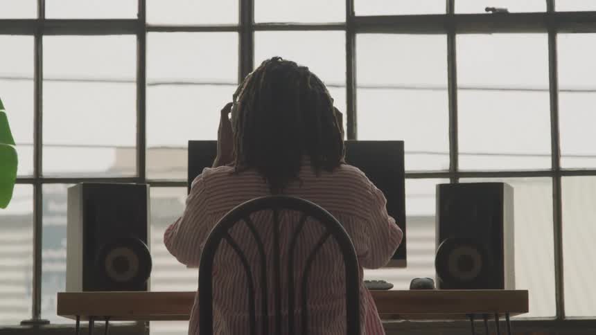 Person sitting at a table in front of a window faces a computer and two speakers while putting on headphones