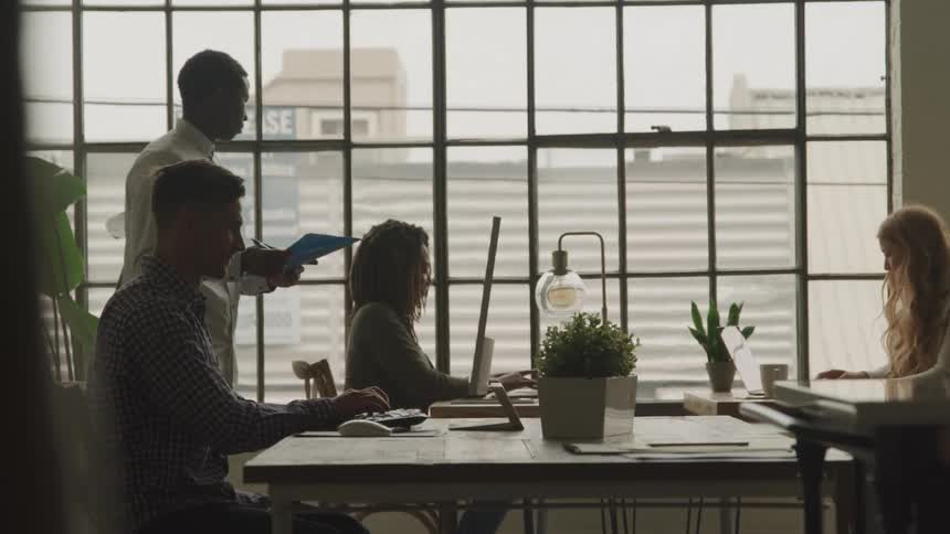 People working in a modern office with one standing and holding a blue folder while others work on computers and laptops