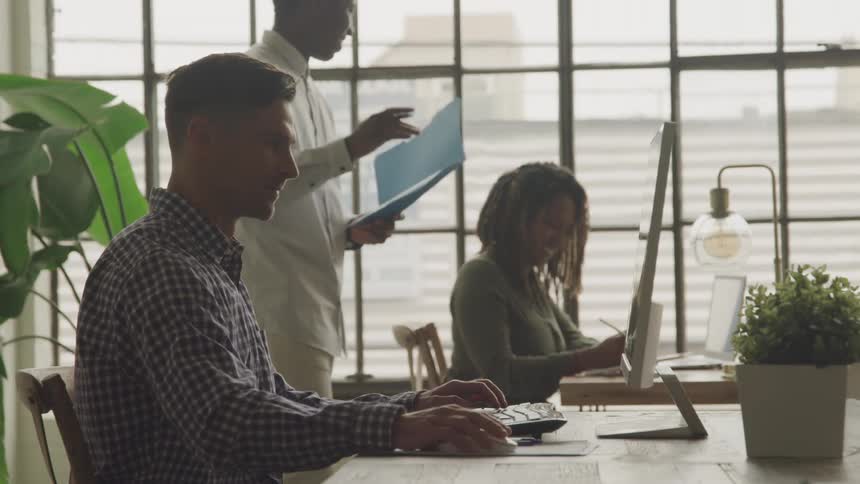A man typing in the office is approached by a coworker