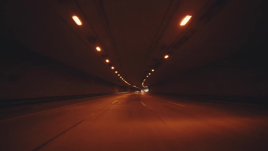 A highway seen from a moving vehicle enters a dimly lit tunnel with its interior stretching forward and rows of lights illuminating the roadway as it emerges on the other side