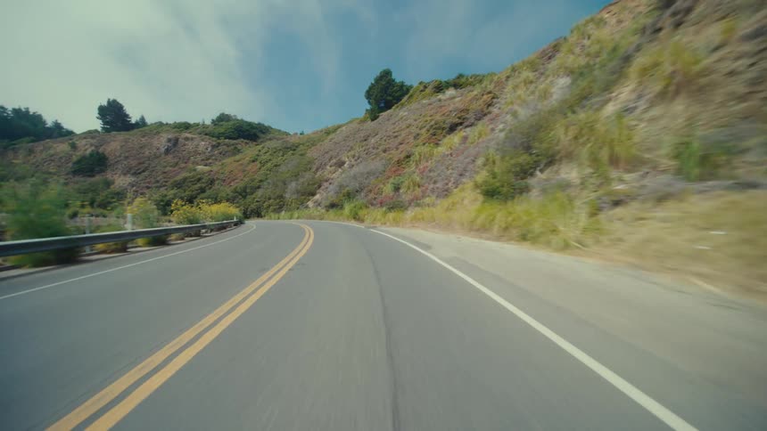 A winding asphalt road with double yellow lines curves along a steep green hillside with a metal guardrail separating the road from a dropoff and a glimpse of the ocean visible in the distance