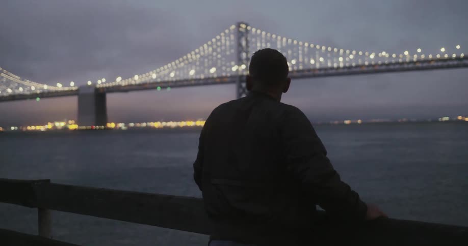 Man stands on a deck at night looking at the golden gate bridge lit up at night