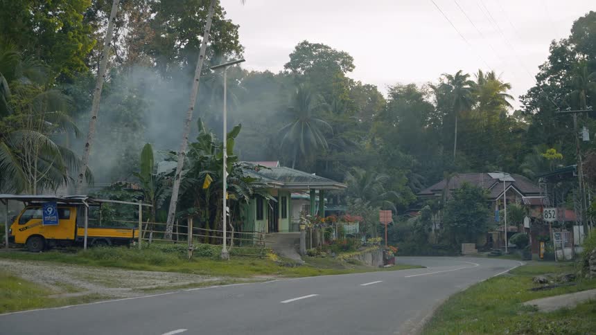 A motorcycle passing a parked yellow vehicle on a rural road lined with houses and palm trees with smoke rising in the background