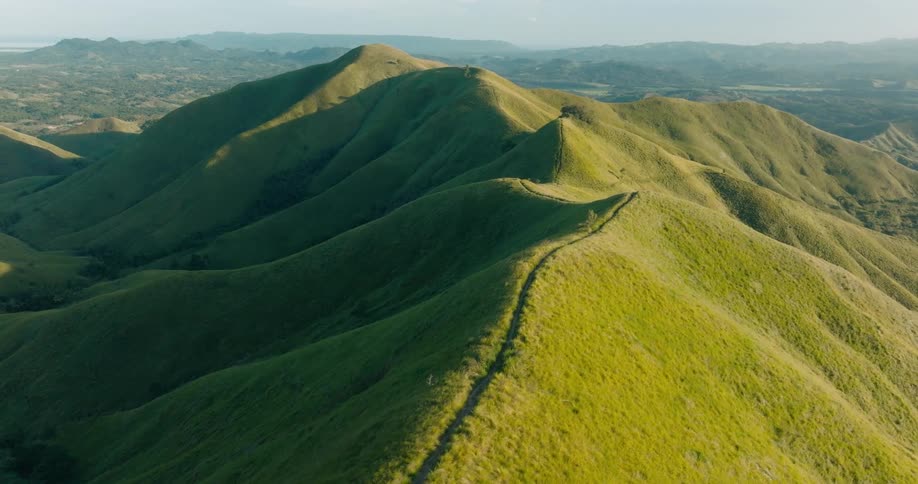 An aerial view shows a series of rolling green hills with a narrow path winding along the crest of one under a hazy sky