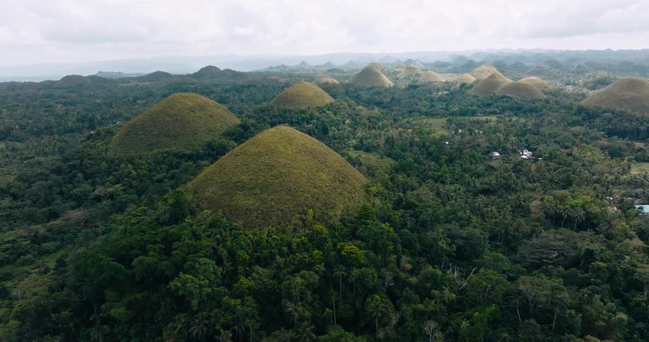 Aerial view moves backward over the iconic chocolate hills rising from a lush tropical forest under a cloudy sky