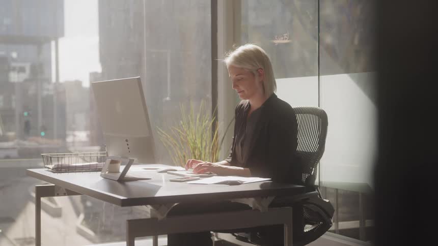 A businesswoman with short lightcolored hair works at a desk in a bright office typing on a computer with a document spread out before her as sunlight streams through the large window behind her