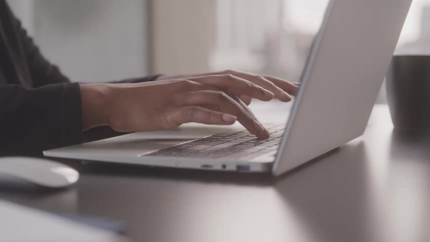Hands typing on a laptop keyboard with a mouse and a mug visible on the side in the office