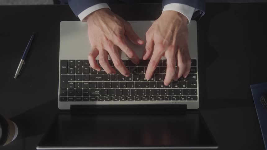 Businessman in a suit types on a laptop keyboard with their hands and the laptop visible from an overhead perspective