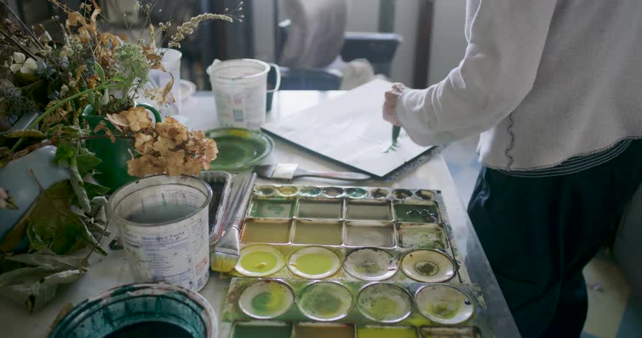 An artist in a studio is working on a painting with a palette of green and yellow paints and various art supplies scattered on the table