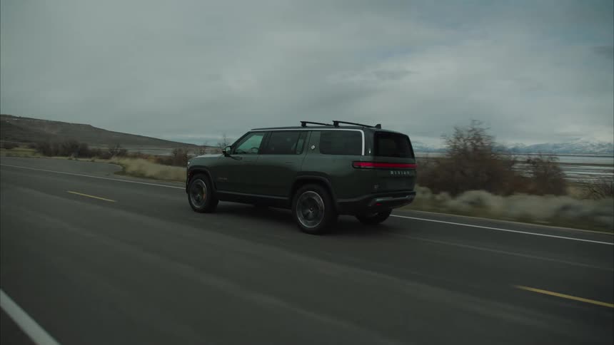 A dark colored rivian r1s suv drives on a paved road through a desolate mountainous landscape under an overcast sky