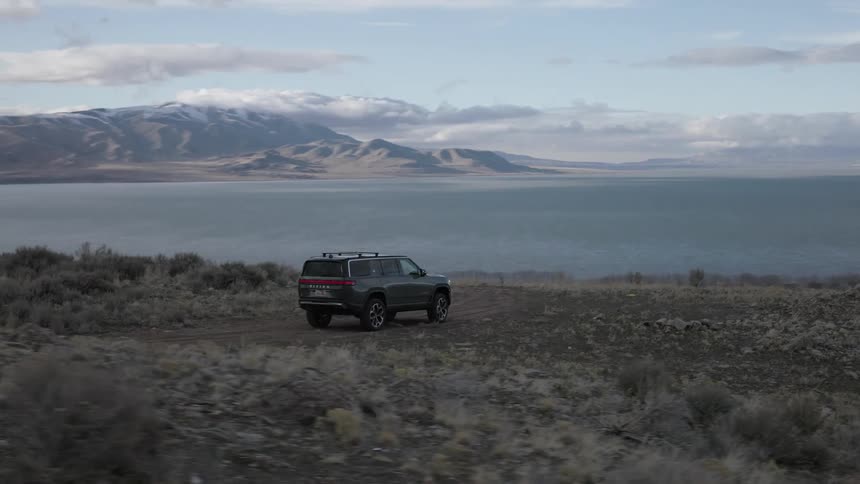 A dark colored rivian r1s suv is parked on a dirt road overlooking a lake and snow capped mountains under a cloudy sky