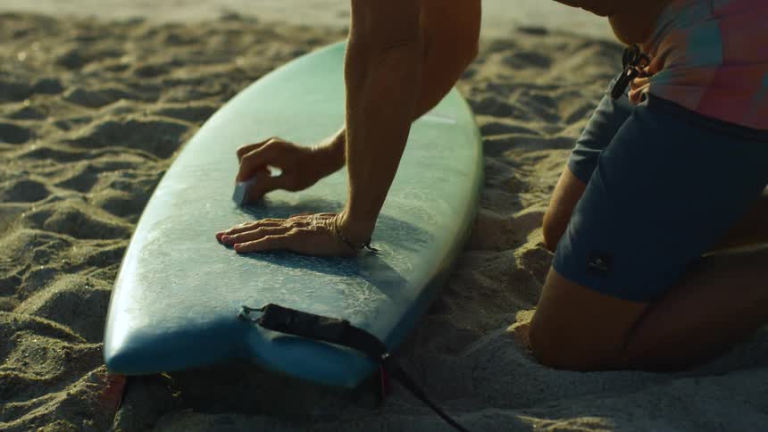 A man is cleaning his surfboard while sitting at the beach