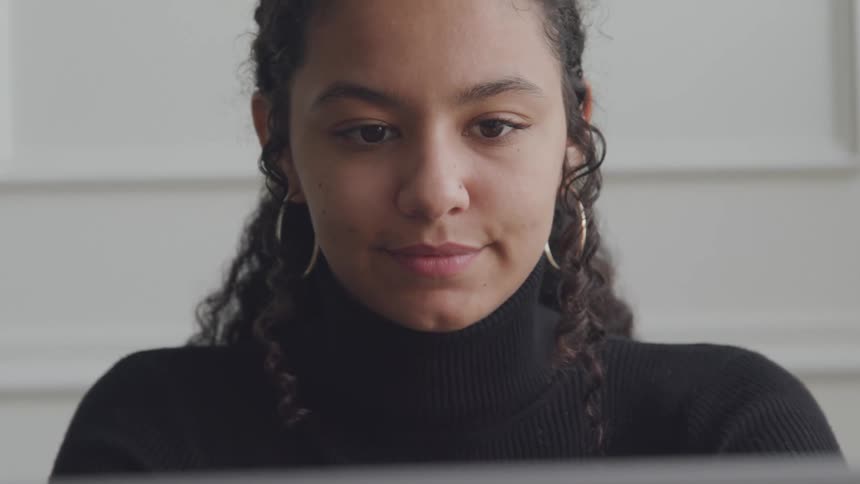 A young woman with curly dark hair and gold hoop earrings wearing a black turtleneck sweater is looking down with a focused expression likely at a screen