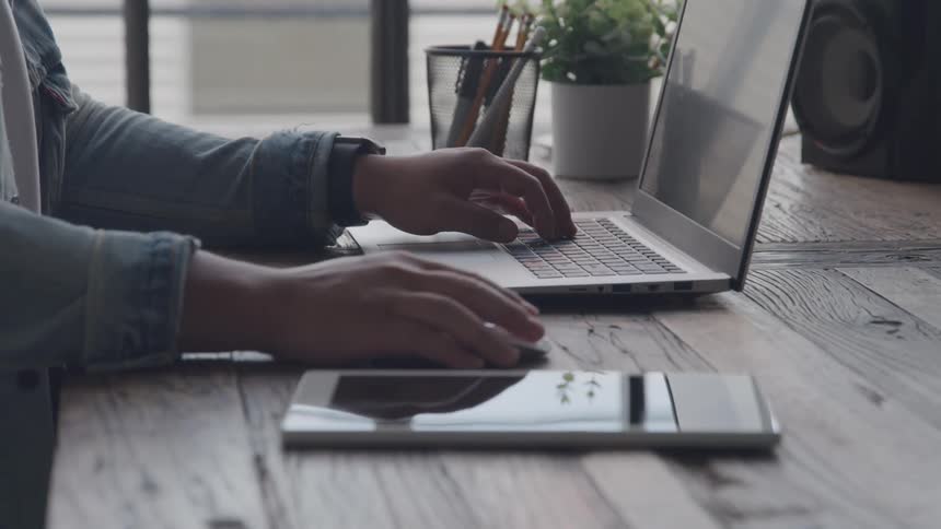 Man sitting at a desk typing on a laptop computer and moving his hand on the mouse near his phone