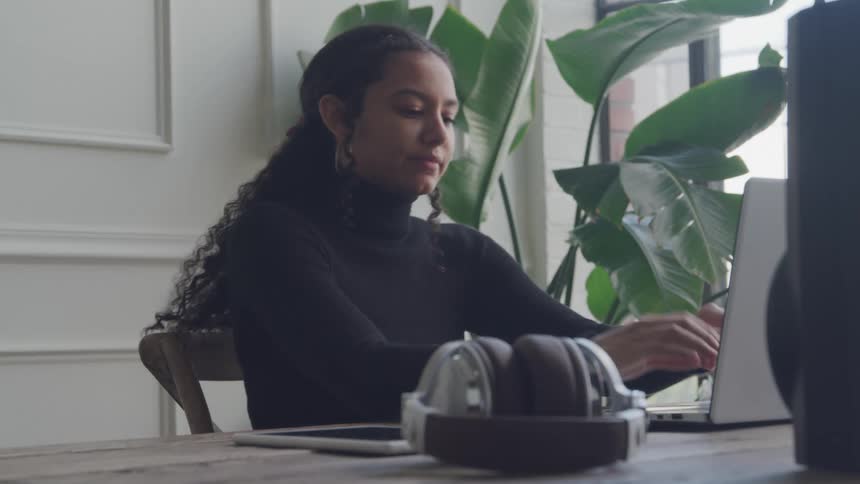 A young woman with a black turtleneck and curly hair is working on a laptop at a wooden table with a pair of headphones a tablet and a large potted plant nearby