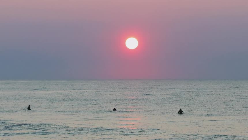 Three people are swimming in the ocean while the sun is setting