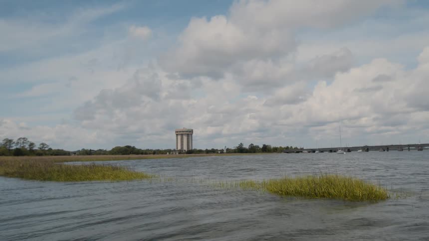A solitary building stands on a flat marshy landscape viewed across rippling water under a partly cloudy sky next to a bridge with traffic