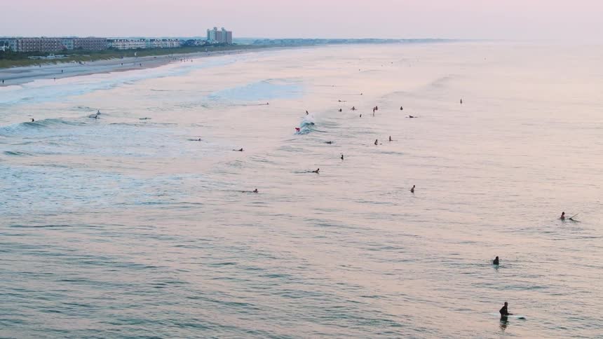 An aerial view shows a coastline with surfers scattered in the ocean small waves rolling in and a city skyline in the distance under a dusky sky
