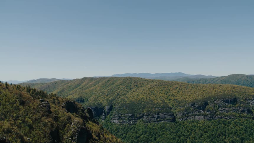 A scenic landscape shot captures a vast mountain range covered in lush greenery under a clear blue sky with rocky cliffs visible in the foreground