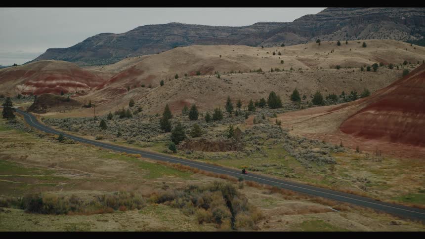 A landscape with distinct geological formations featuring a prominent reddishbrown layered hill surrounded by more muted tan and green vegetation and a road winding through the scene where a biker is riding a motorcycle under a daytime sky