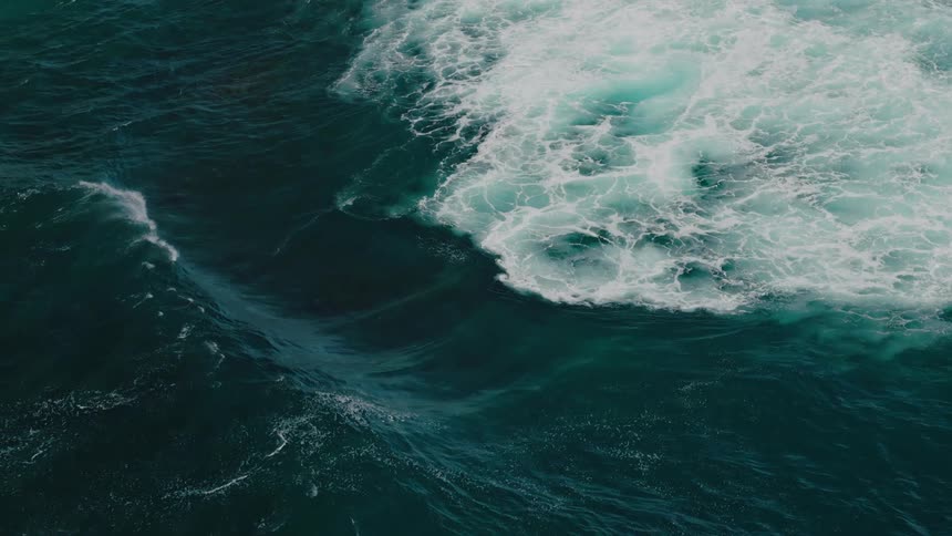 An aerial view of a large wave crashing into the ocean creating a swirl of white foam against the deep blue water with smaller waves and splashes visible around it