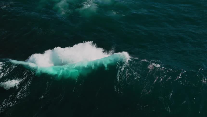 An aerial view shows a large wave crashing into the deep blue ocean creating a burst of white foam and a turquoise splash against the dark water