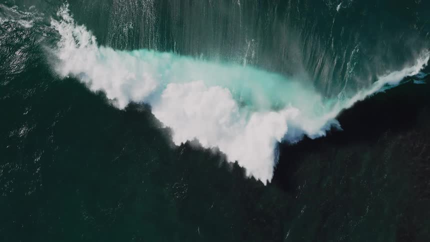 An aerial view of a large wave crashing into the ocean creating a burst of white foam and a turquoise splash against the dark water