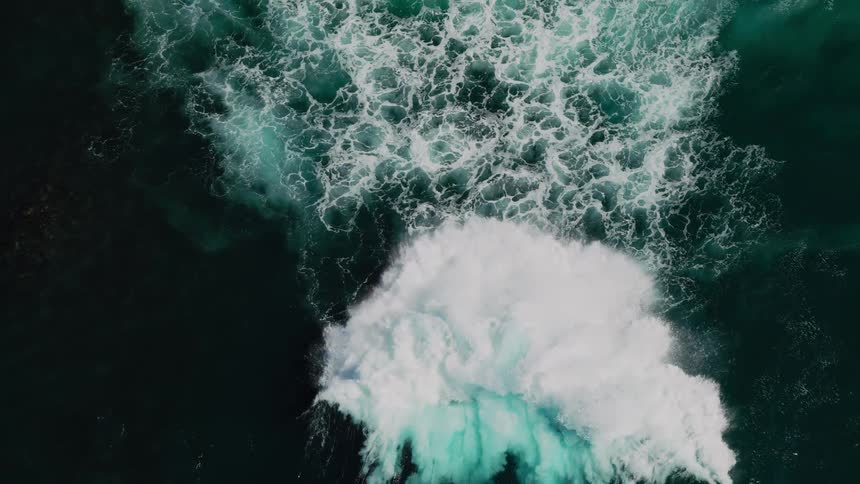 A large wave crashing into the ocean creating a burst of white foam and turquoise water against the dark sea