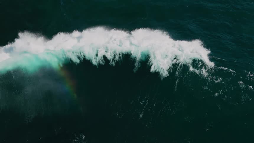 An aerial view shows a large wave breaking in the ocean with white foam and a faint rainbow visible against the deep blue water