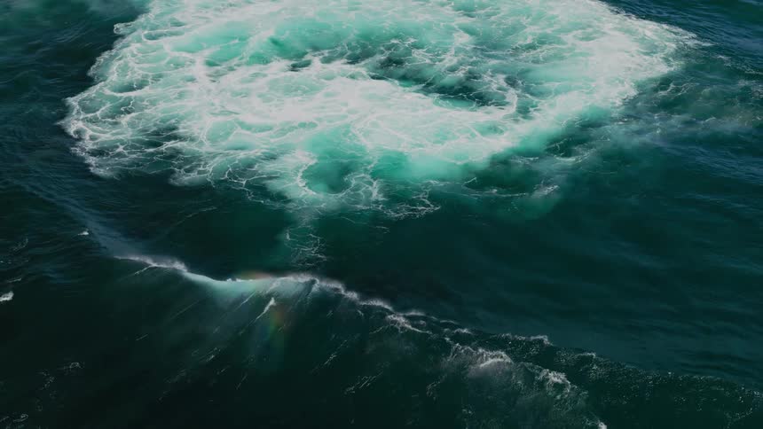An aerial view shows a wave crashing into the ocean creating a swirl of white foam and turquoise water against the dark sea