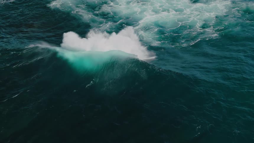An aerial view shows a turbulent sea with white foam churning against the deep blue green water