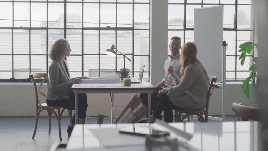 Two people sit in an office engaging in a conversation with a woman in a blazer in a bright room with a large window