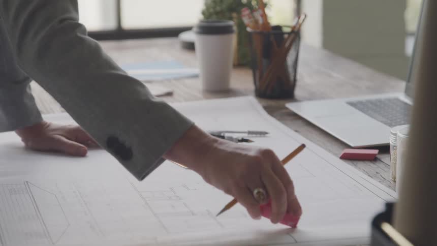 A woman erasing something on architectural plans she is holding a pencil as she uses a pink eraser to erase on the paper