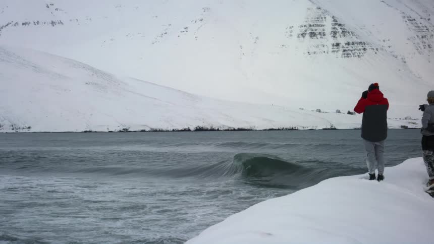 Two individuals stand on a snowcovered bank overlooking a body of water near a large snowcovered landmass