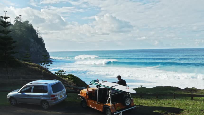 Man standing near the coast looks towards the surfboard on a nearby car on a sunny daytime