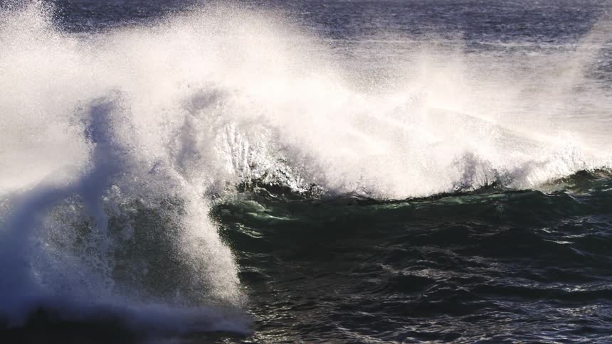 A rolling wave in the ocean its peak topped with white spray against the backdrop of a sunlit sparkling sea